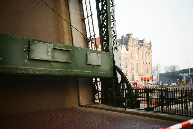 The image features an urban scene with a focus on an old, green, metal bridge or overpass that spans above what appears to be a street. To the left of the frame, there is a large building, possibly a church due to its architectural style and the presence of a tall tower. This structure has intricate designs and details, suggesting it could be historic in nature. The sky overhead is overcast, indicating a gloomy or cloudy day. In the foreground, the lower part of a traffic signal is visible along with a roadside railing, giving a sense of depth to the image.
