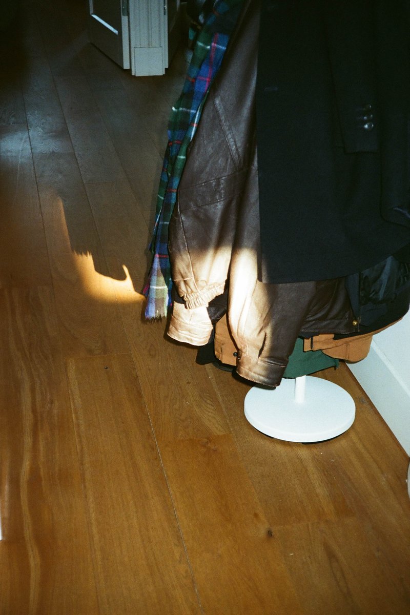 The image shows an indoor setting with various clothing items hanging on a rack against a wall. In the foreground, there's a white object that looks like a piece of furniture or household item. The floor appears to be made of wood and the walls have a simple design.