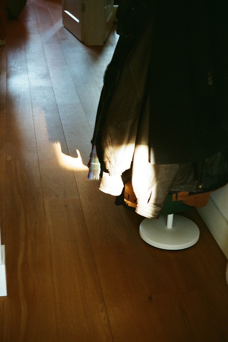 The image shows an indoor setting with a long, straight view down a hallway. At the end of the hallway, there's a bright light source, possibly sunlight, creating a visible ray of light on the floor. To the left side of the image, a series of hanging clothes are seen, indicating that this could be a dressing room or a closet. The colors in the image are muted, with a wooden floor and walls that are not highly detailed. There's a sense of depth due to the perspective view down the hallway.