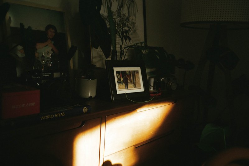 The image depicts an interior space with natural light filtering through a gap in window blinds or curtains. Sunlight is creating shadows that play on the window, possibly indicating it is morning or afternoon when the sun's rays are at their strongest. To the right, there appears to be a photograph of a person in a frame, though the details of the photo are not clear due to the image's resolution and size. The setting suggests a home environment with personal items such as a book and a small plant or vase visible on a surface, which could be a desk or side table.