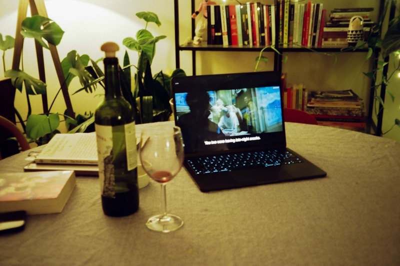 The image shows an indoor setting with a table displaying a laptop computer. The laptop screen is open, but the content displayed is not legible in this description. To the right of the laptop, there is a wine glass and a bottle, suggesting a relaxed or social environment. In the background, there is a bookshelf filled with various items, including what appears to be a collection of wine glasses, books, and other decorative objects. The overall scene suggests a cozy home environment, possibly used for work or leisure activities. There is no text visible in this image.