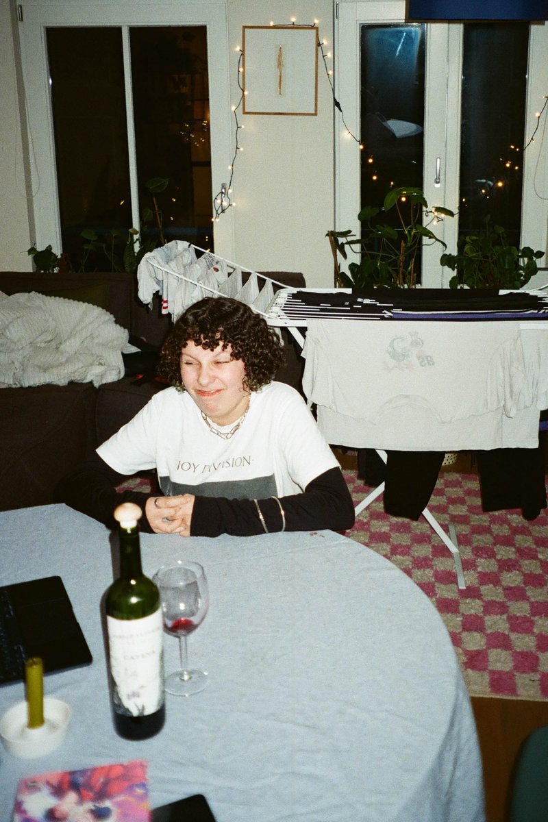 The image shows an indoor scene with a young woman seated at a dining table in front of a window decorated with lights. She appears to be engaged in some activity on the table, which is covered with various items including a wine bottle and a glass. A potted plant can be seen near the window.