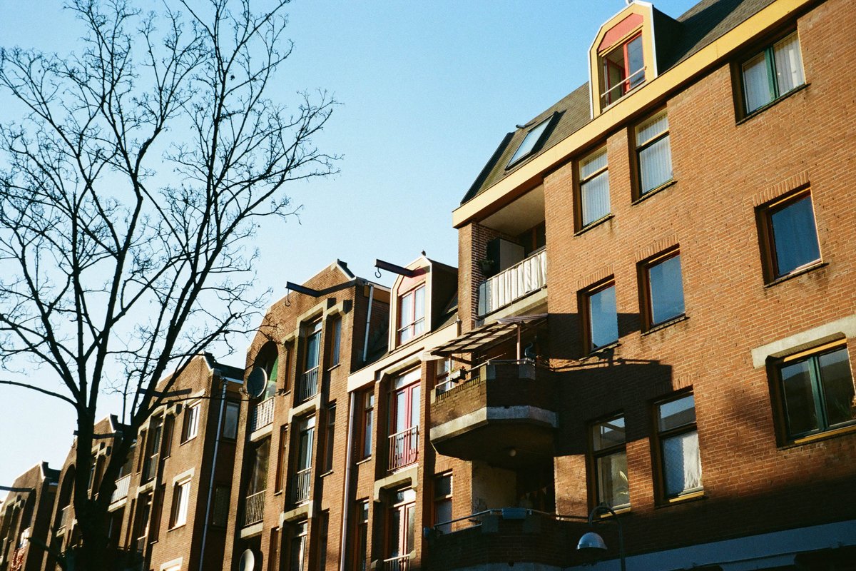  The image depicts an urban street scene. There are several buildings lining the street, which appear to be residential apartments with balconies. It is daytime, and there's no visible snow or ice. There are trees that add some greenery to the scene. 