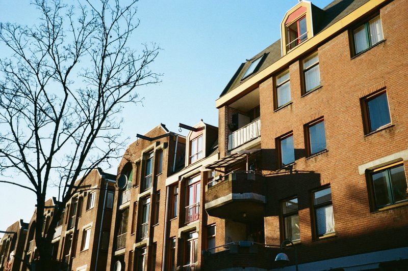 The image depicts an urban street scene. There are several buildings lining the street, which appear to be residential apartments with balconies. It is daytime, and there's no visible snow or ice. There are trees that add some greenery to the scene.