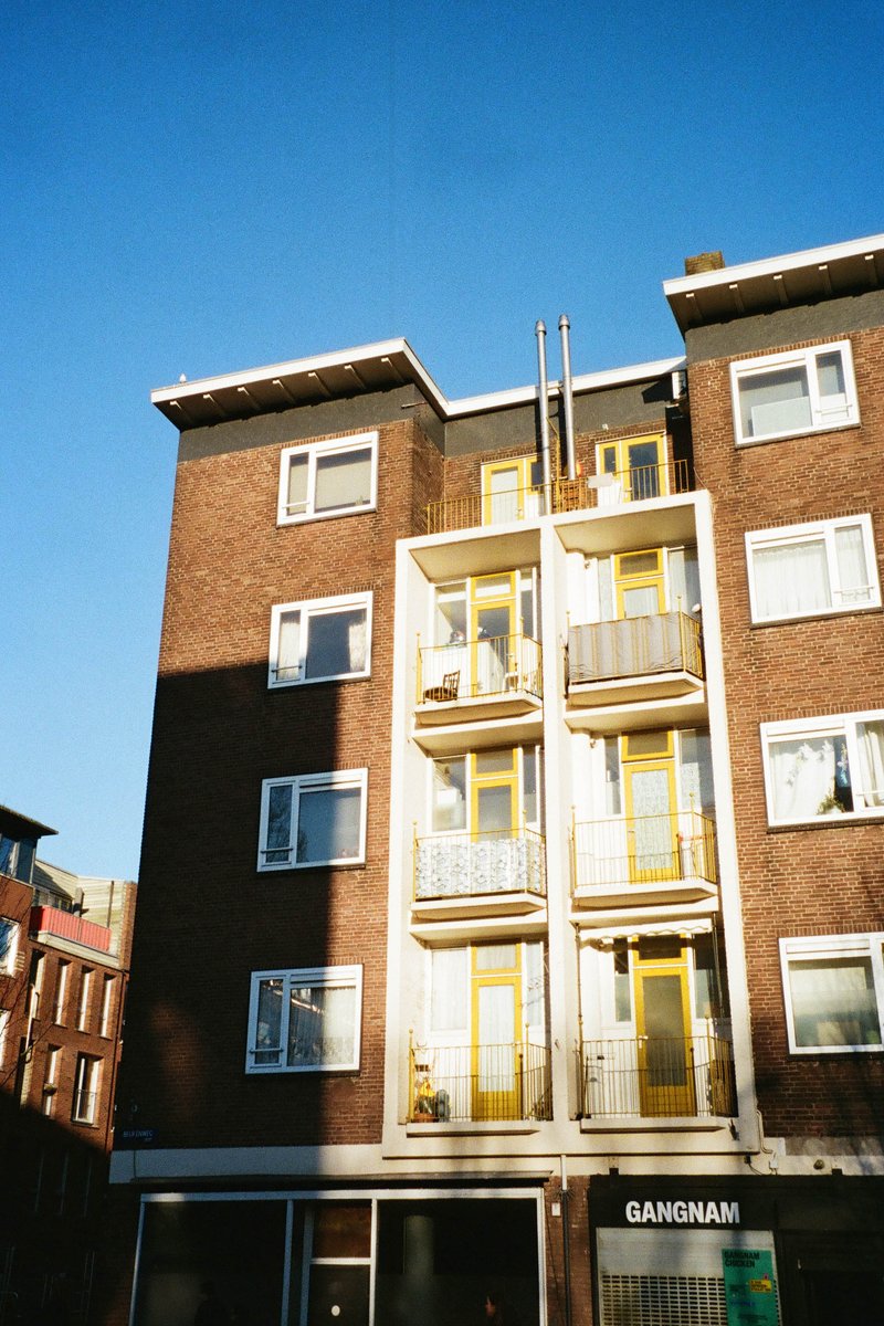 The image shows a multi-story building with multiple windows of various sizes. The building appears to be residential or commercial with balconies visible on some floors. The facade is painted in a light color, possibly yellow, with a darker trim around the edges of the windows and balconies. There are no visible texts or distinctive branding. The sky is clear blue, suggesting it may be a bright day without any significant cloud cover.