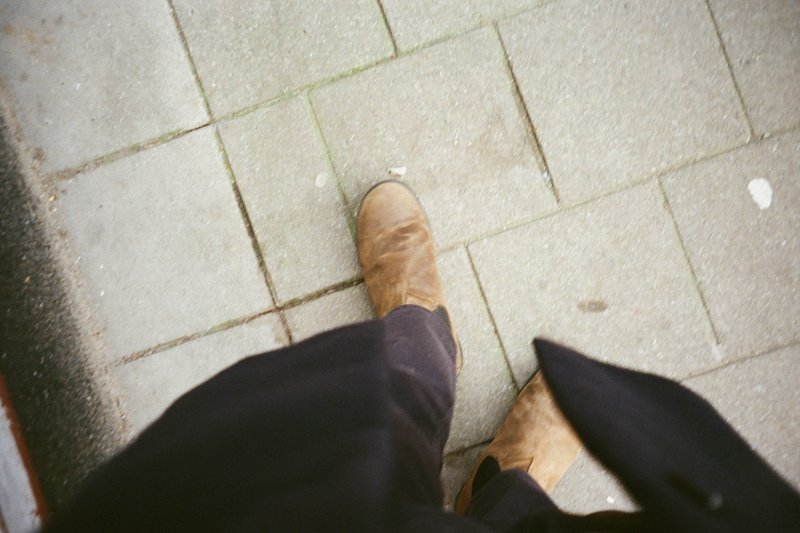 The image captures a person walking down a cobblestone street lined with brick buildings. The individual is dressed in dark clothing and boots, with one foot slightly ahead of the other. The sidewalk and steps are visible, creating a sense of depth and perspective.