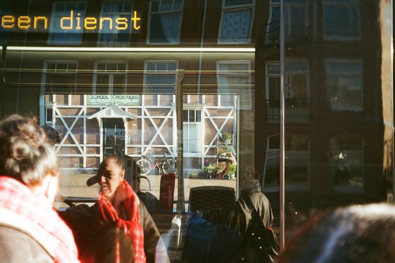 The image shows a scene from a busy street with people walking by. There's a storefront displaying various items, possibly merchandise or decorations. An individual is taking a photo of the scene outside through the shop window. The weather appears to be overcast, suggesting it might be raining or about to rain.