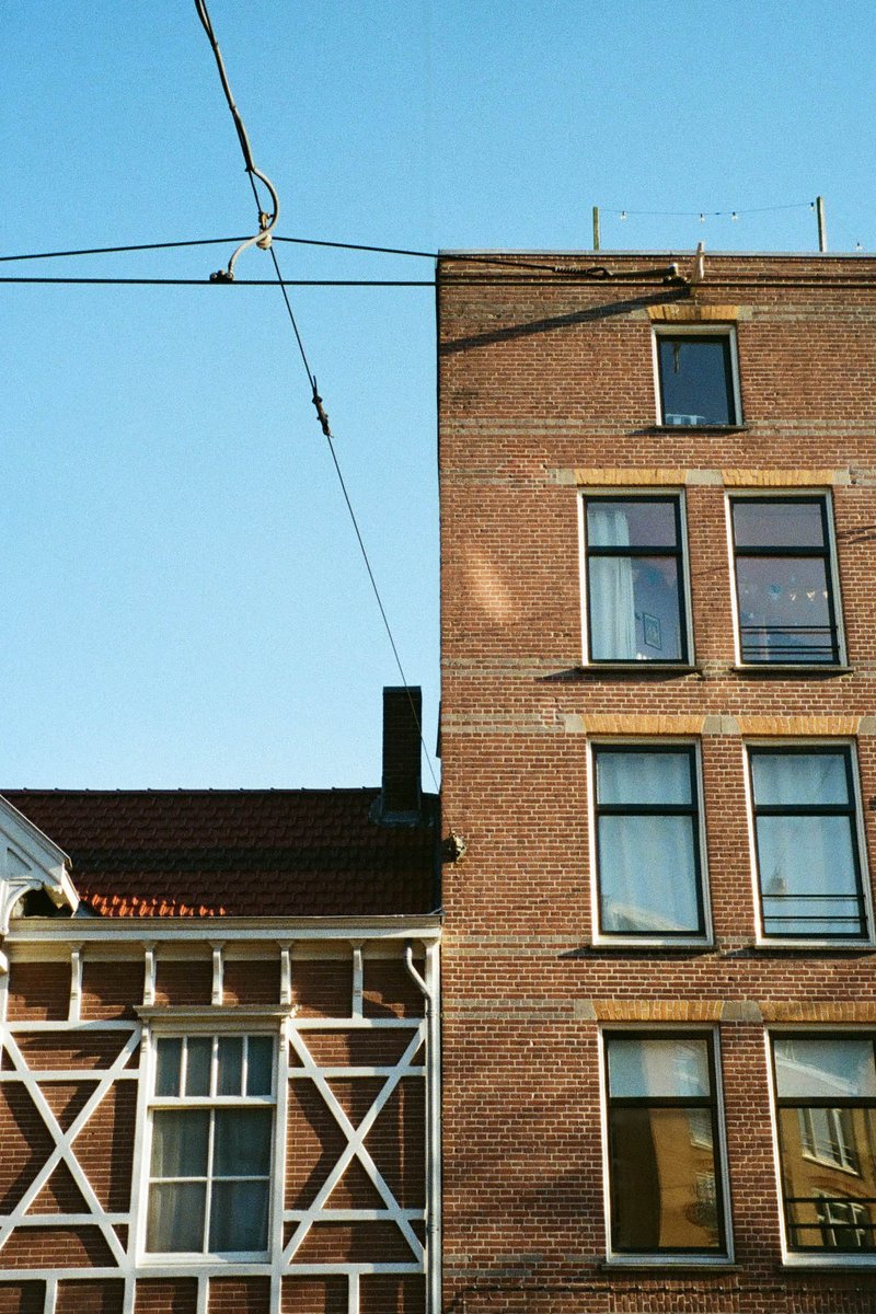 The image shows a view of two buildings. On the left is an old, tall building with a distinct red brick exterior. To its right is another building that appears to be older with multiple windows. Both structures have different architectural features and colors, with the left one being more prominent due to size and location in the frame. The image also captures a clear sky above and a street sign below.