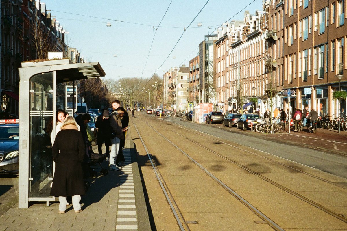  The image captures an urban scene at an intersection. There is a tram line with a train car stationed at a stop. People are waiting for the train, standing on the platform near the tracks. In the background, there are buildings with a European architectural style, including those with balconies. The sky above is overcast, suggesting it might be a cloudy day. 