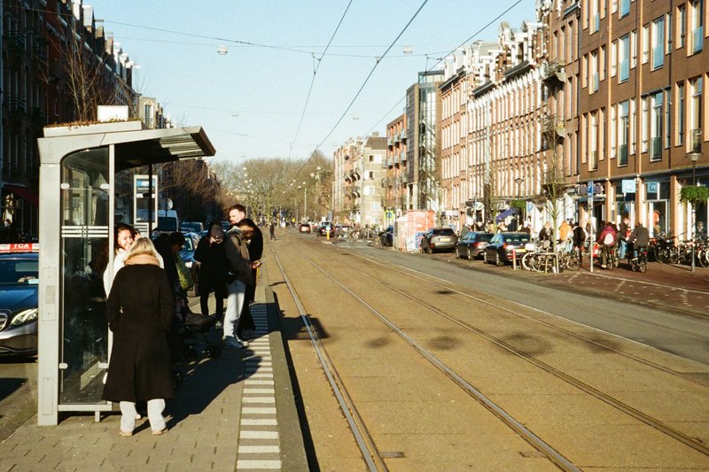 The image captures an urban scene at an intersection. There is a tram line with a train car stationed at a stop. People are waiting for the train, standing on the platform near the tracks. In the background, there are buildings with a European architectural style, including those with balconies. The sky above is overcast, suggesting it might be a cloudy day.