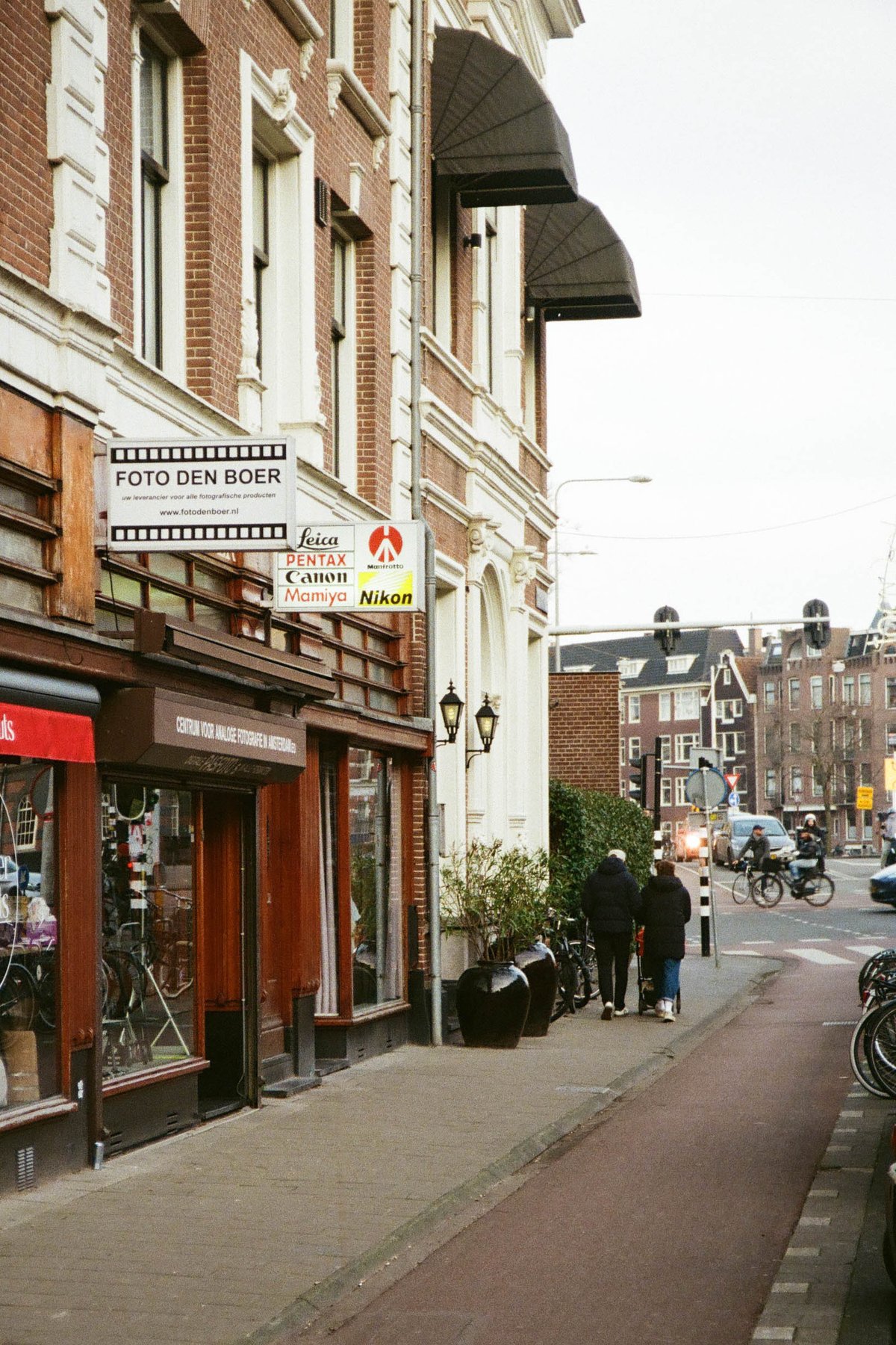 The image depicts a city street scene, with an old European-style building on the left side of the frame, featuring a sign that reads "Bistro." On the right, there's a modern establishment with a red and white logo. A pedestrian is walking on the sidewalk in front of these shops, and the buildings are adjacent to one another, giving the impression of a quaint city neighborhood. 