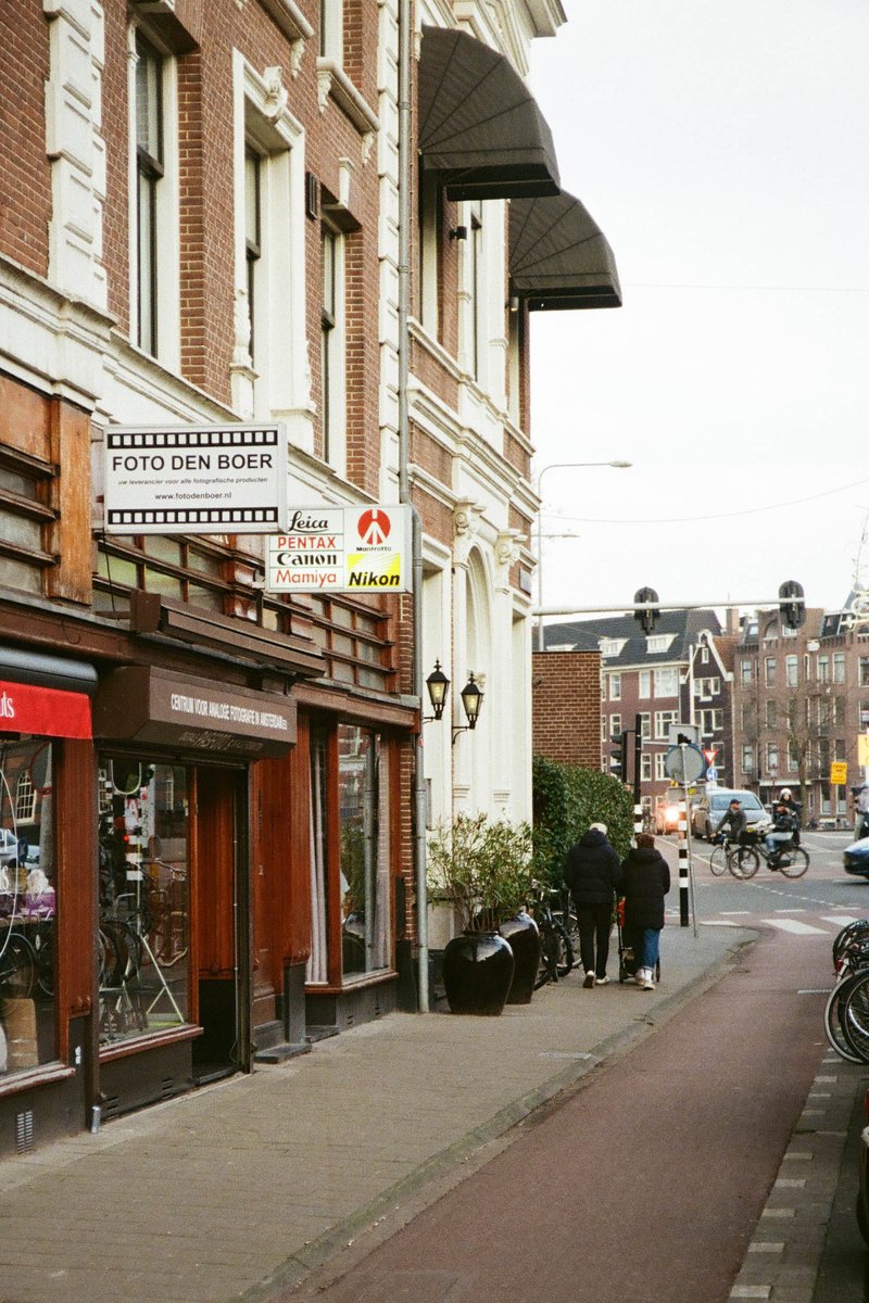 The image depicts a city street scene, with an old European-style building on the left side of the frame, featuring a sign that reads "Bistro." On the right, there's a modern establishment with a red and white logo. A pedestrian is walking on the sidewalk in front of these shops, and the buildings are adjacent to one another, giving the impression of a quaint city neighborhood.