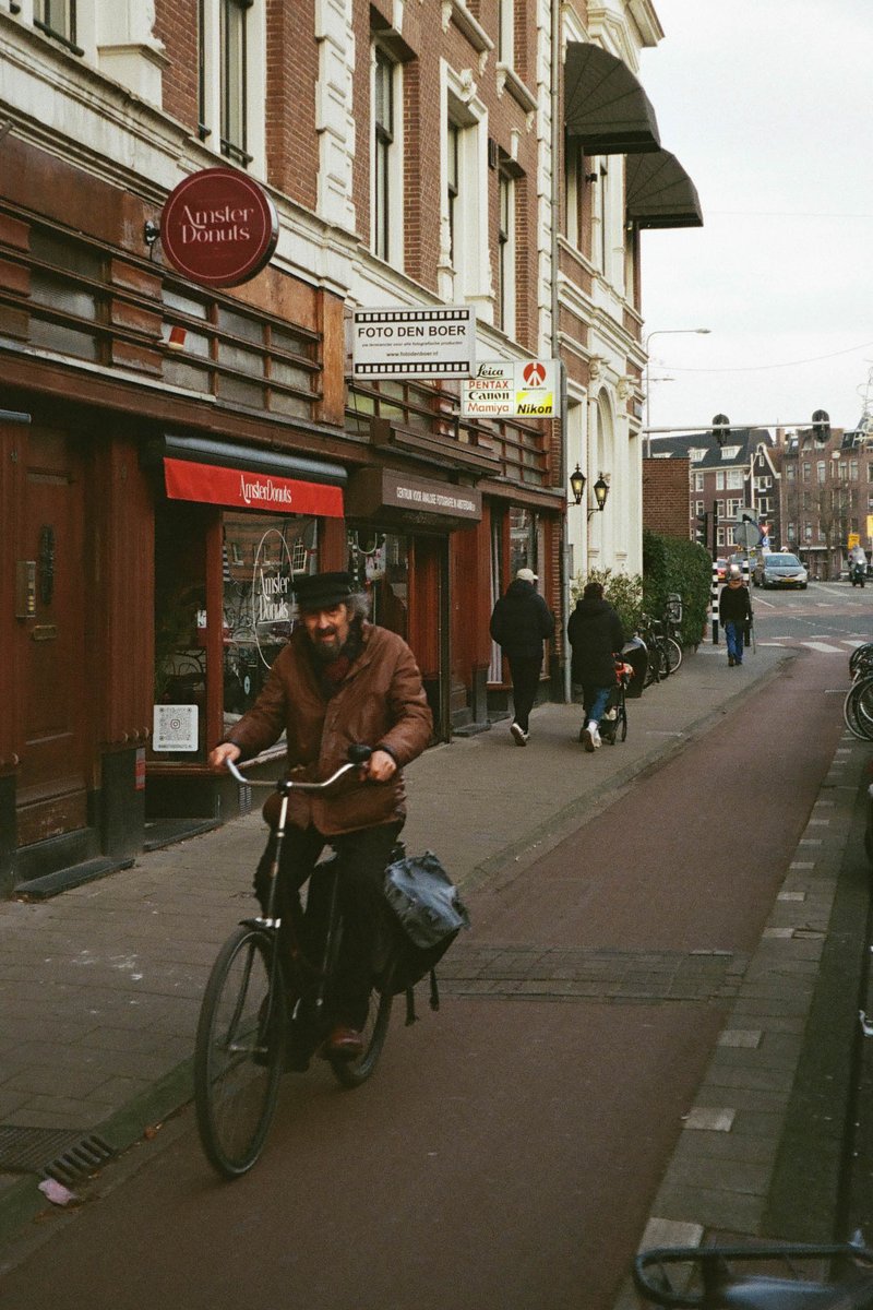 The image depicts an outdoor urban scene with a man riding a bicycle down a bike lane. He is wearing casual clothing and appears to be carrying a bag or pack. There are buildings on either side of the road, with signage indicating the presence of businesses, such as a cafe and a shop. The photo has a vintage look due to its grainy texture and dated feel.