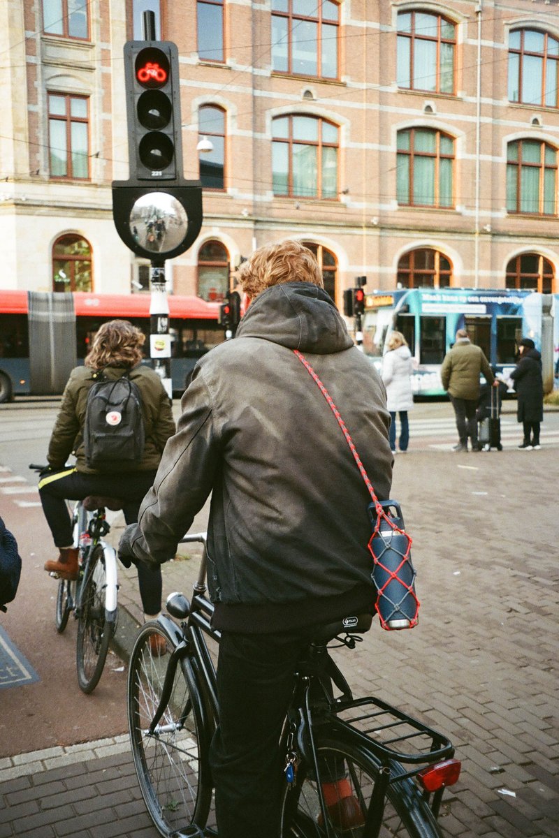 In the center of the image, two men are riding bicycles on a bustling city street. They are wearing backpacks and are positioned under traffic lights. The backdrop features a row of buildings with a mix of brick and glass facades. The atmosphere suggests an urban setting during daylight hours.