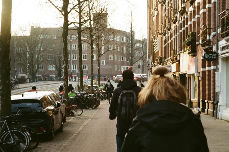 The image captures a vibrant street scene. Pedestrians can be seen walking along the sidewalk, while bicycles are parked on the side. Buildings line both sides of the street, and there's lush greenery scattered around. It's a typical day in this city area, with people going about their daily routines.