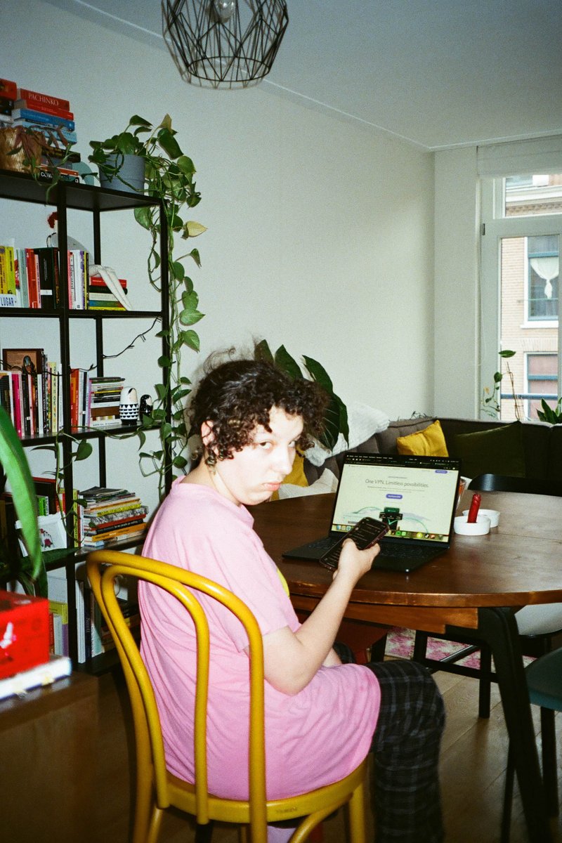 The image shows a cozy room with a woman sitting at a dining table using a laptop. She is wearing a pink top and looking down at the screen of her laptop. To her right, there is a bookshelf filled with various items. Behind her, part of a plant can be seen, adding a touch of greenery to the scene. The room appears well-lit and comfortable, suggesting a personal space where the woman might be working or browsing online.