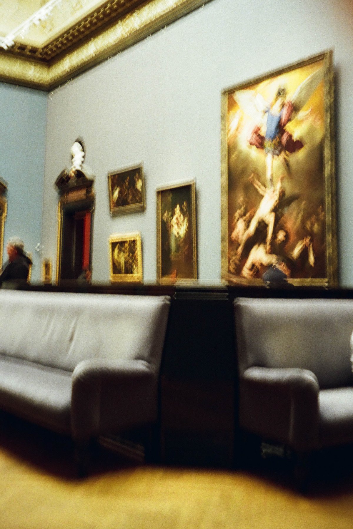  This is a photograph capturing an interior scene of a museum. The focal point is a room filled with art, featuring large paintings displayed on walls. In the foreground, there are two benches placed next to each other on the floor, inviting visitors to sit and admire the artwork. A solitary person can be seen in the background, adding a human element to the scene. The ceiling of the room has intricate patterns and decorations, which contribute to the grandeur of the space. 