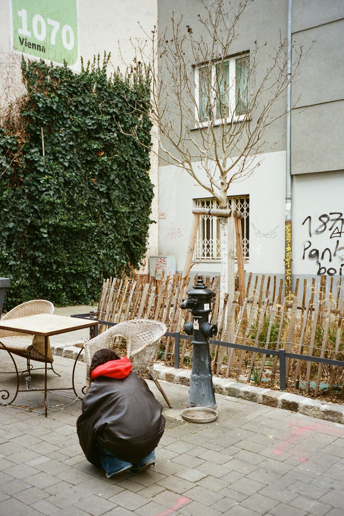  The image captures an urban scene featuring a street corner with a metal fire hydrant in the foreground. A person is seated on a curb adjacent to a tree and a small table, likely engaged in a conversation or waiting for someone. In the background, there's a building with signage and graffiti on its exterior walls. The atmosphere appears calm and somewhat solitary. 