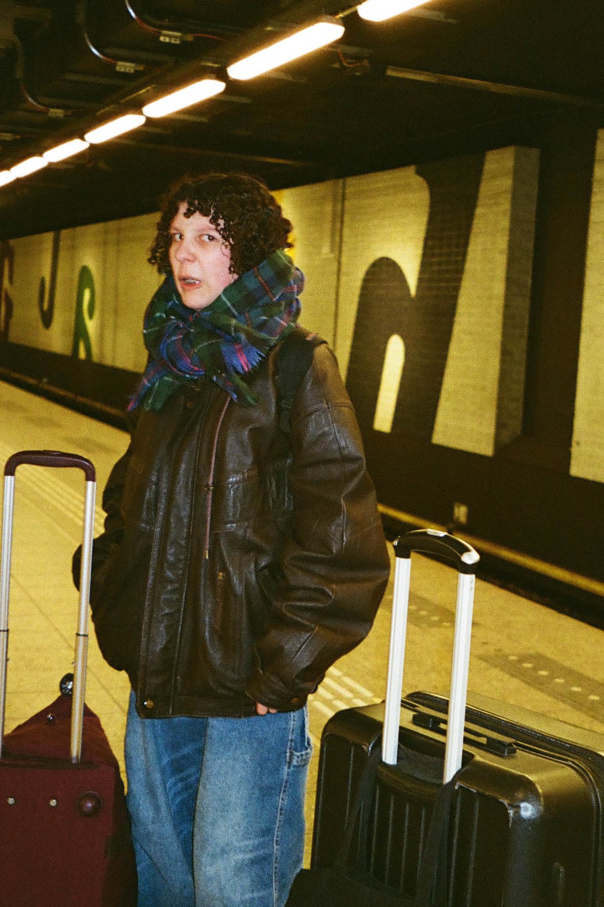 The image features an individual standing at a subway station platform with luggage nearby. They are wearing casual clothing, including a jacket, and appear to be waiting for a train. The person is facing the camera, and the background shows a train on the track and a wall with advertisements. The colors in the image include shades of blue, red, green, and various hues in the individual's outfit and the luggage. 