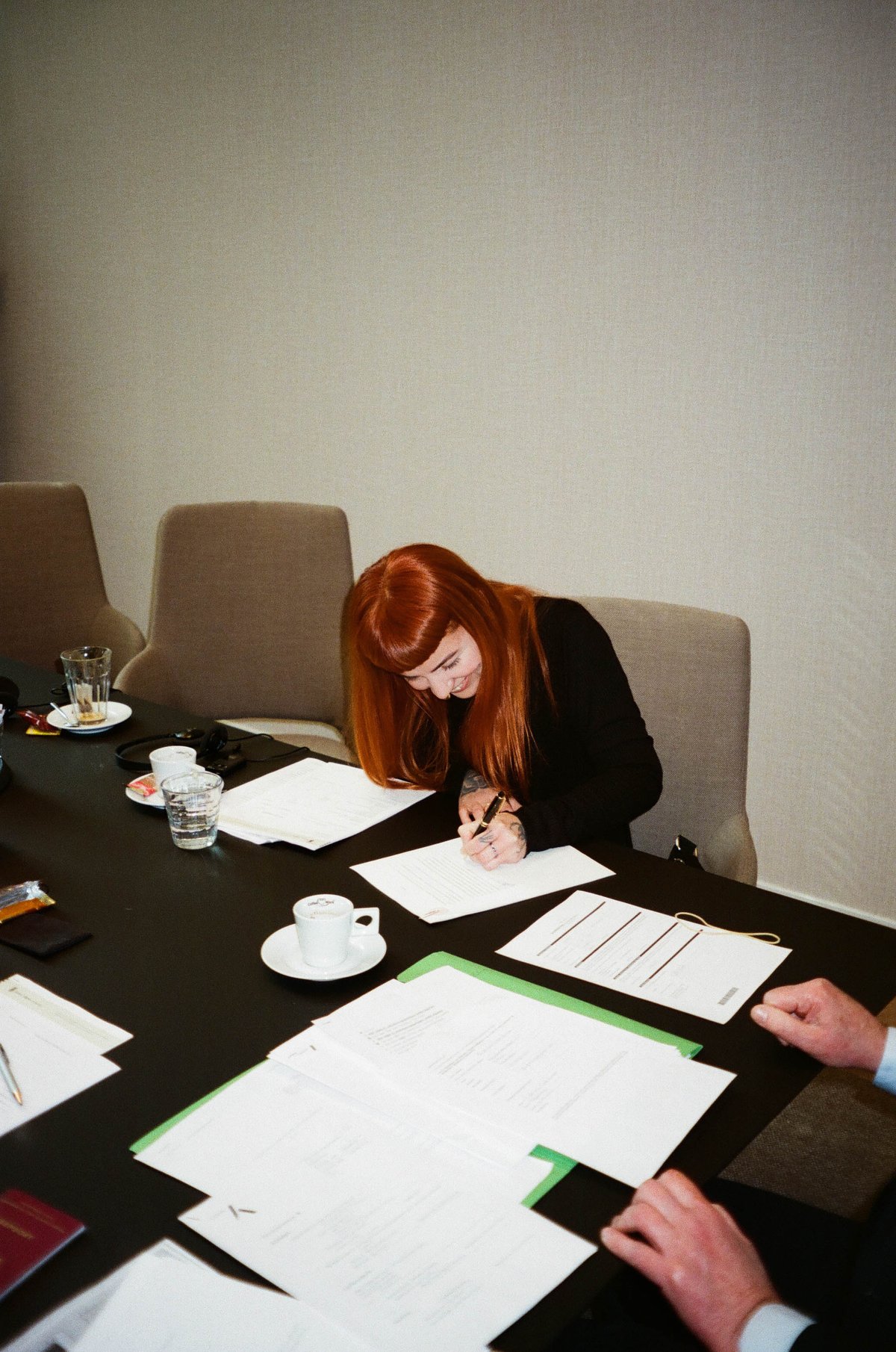  The image shows an indoor setting with a woman sitting at a table, writing or signing papers. She appears to be in a professional environment, given the presence of other individuals and documents on the table. There are two other people visible behind her; one is seated while the other stands. The room is well-lit, and there's a sense of formality due to the attire and setting. 
