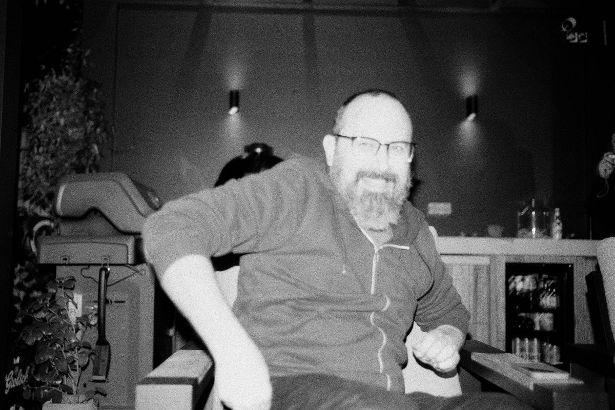  The image is quite blurry, but it appears to be a black and white photograph of a man sitting at a table with his head tilted slightly to one side. His expression seems contemplative or relaxed, and he's wearing glasses. Behind him, there is a room with a warm ambiance, possibly a home setting with various objects on shelves and tables, including what looks like bottles. The overall mood of the image is somewhat mysterious due to the graininess and the low light conditions. 