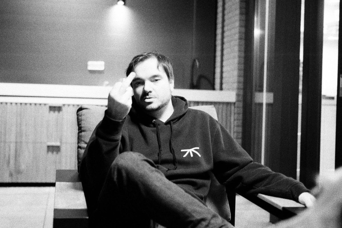  The image shows a man seated with his hand resting on his chin, appearing thoughtful or contemplative. He is indoors, surrounded by a darker environment. There's a chair visible behind him, and the setting seems casual. 